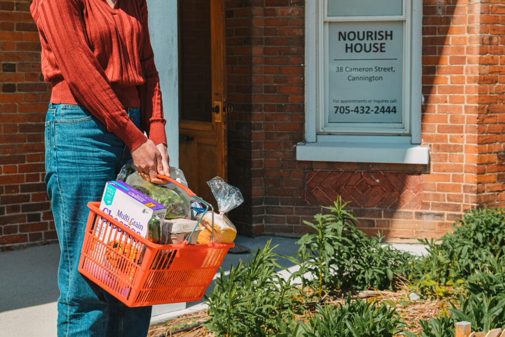 A person holding a basket of groceries in front of Nourish House Food Bank