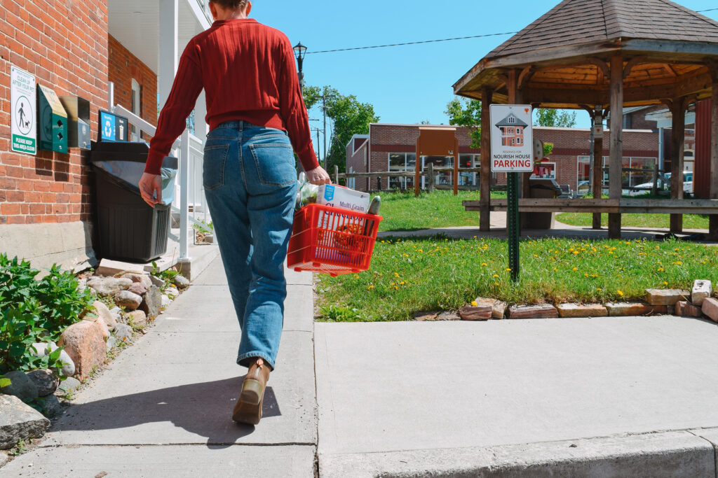 A woman with a full basket of groceries walks up a path toward Nourish House Food Bank