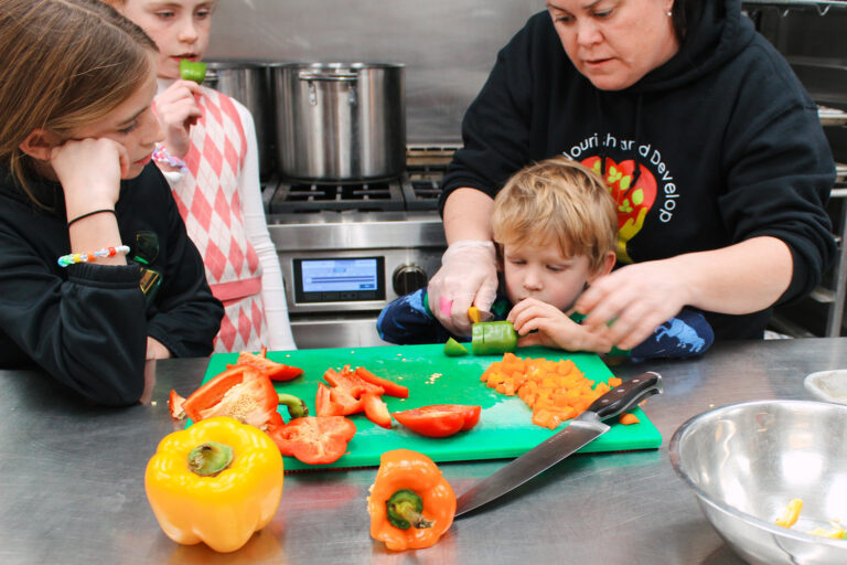 An adult helps a child cut a green pepper during a cooking class