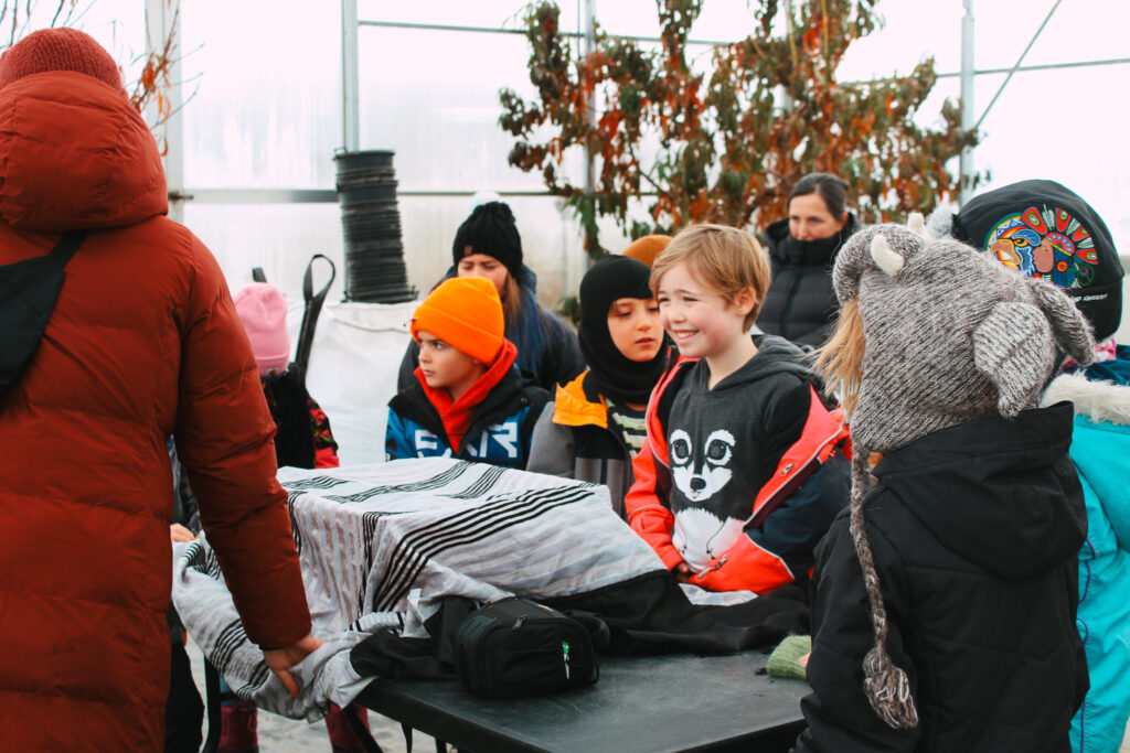 Students in a greenhouse watch eagerly for the instructor to reveal what is under a cloth during one of our food literacy camps.