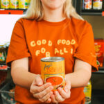 A staff member holds up a can of soup while wearing a shirt that says 'Good Food for All'