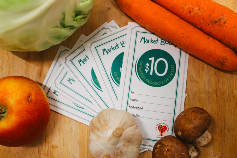 Market Bucks (Mobile Food Market gift cards) on a wooden cutting board surrounded by vegetables.
