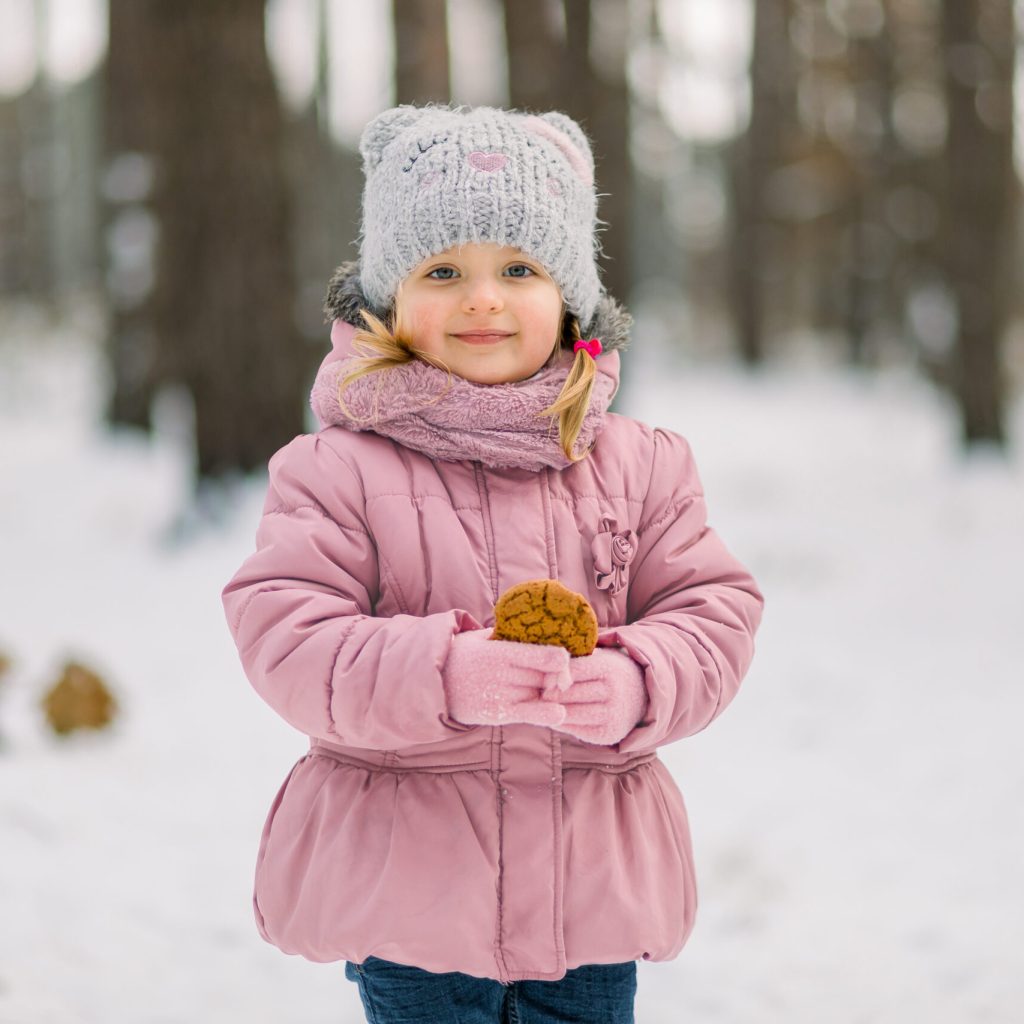 Portrait of little child girl, wearing pink warm coat and gray cap, posing on fresh air in winter snowy forest with tasty homemade cookie in hands.